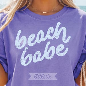 May include: A lilac-colored t-shirt with the words "beach babe" in a distressed, light blue font. The shirt also features the brand name "littlstix" in a banner at the bottom.