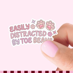 May include: A die-cut sticker featuring the text EASILY DISTRACTED BY TOE BEANS in a rounded pink and purple font, accompanied by two light pink cat paw prints, two small flowers, and two four-pointed star icons on a white background.