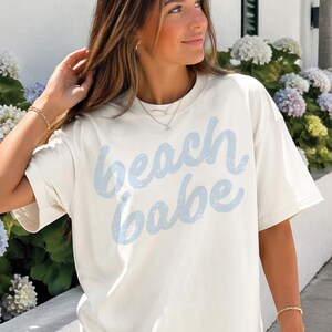May include: Cream-colored t-shirt with the words "beach babe" in light blue script. The shirt has short sleeves and a relaxed fit. The text has a distressed, vintage look.