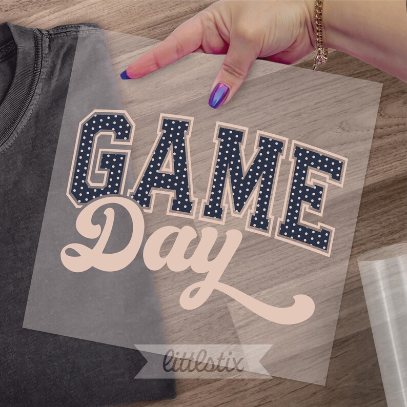 Op de afbeelding: Een hitteoverdrachtssticker met de tekst GAME DAY. Het woord GAME is in een donker marineblauw bloklettertype met witte stippen en een lichtbeige omlijning. Het woord Day is in een lichtbeige cursief lettertype op een transparant vel.