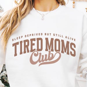 Puede incluir: Sudadera blanca con el texto "SLEEP DEPRIVED BUT STILL ALIVE" encima de "TIRED MOMS Club" en letras marrones. La sudadera tiene cuello redondo y mangas largas. La imagen muestra la parte superior del cuerpo de una persona que lleva la sudadera.