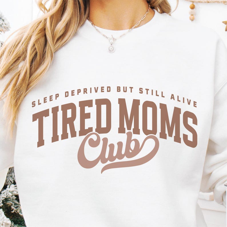 Puede incluir: Sudadera blanca con el texto "SLEEP DEPRIVED BUT STILL ALIVE" encima de "TIRED MOMS Club" en letras marrones. La sudadera tiene cuello redondo y mangas largas. La imagen muestra la parte superior del cuerpo de una persona que lleva la sudadera.