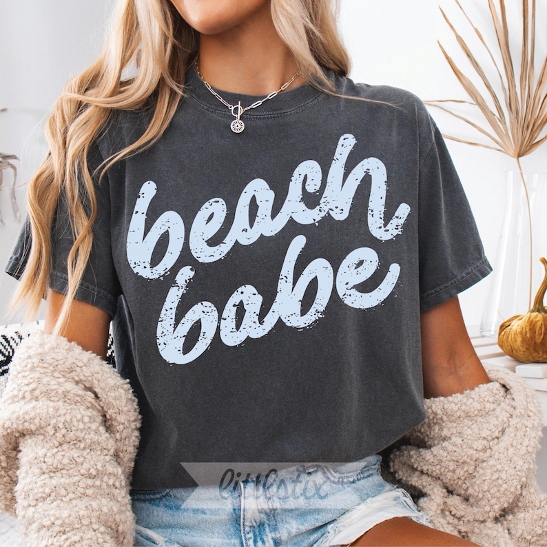 May include: Dark gray t-shirt with the words "beach babe" in light blue script. The shirt has a vintage, washed look. The model is wearing denim shorts and a cream-colored sweater.