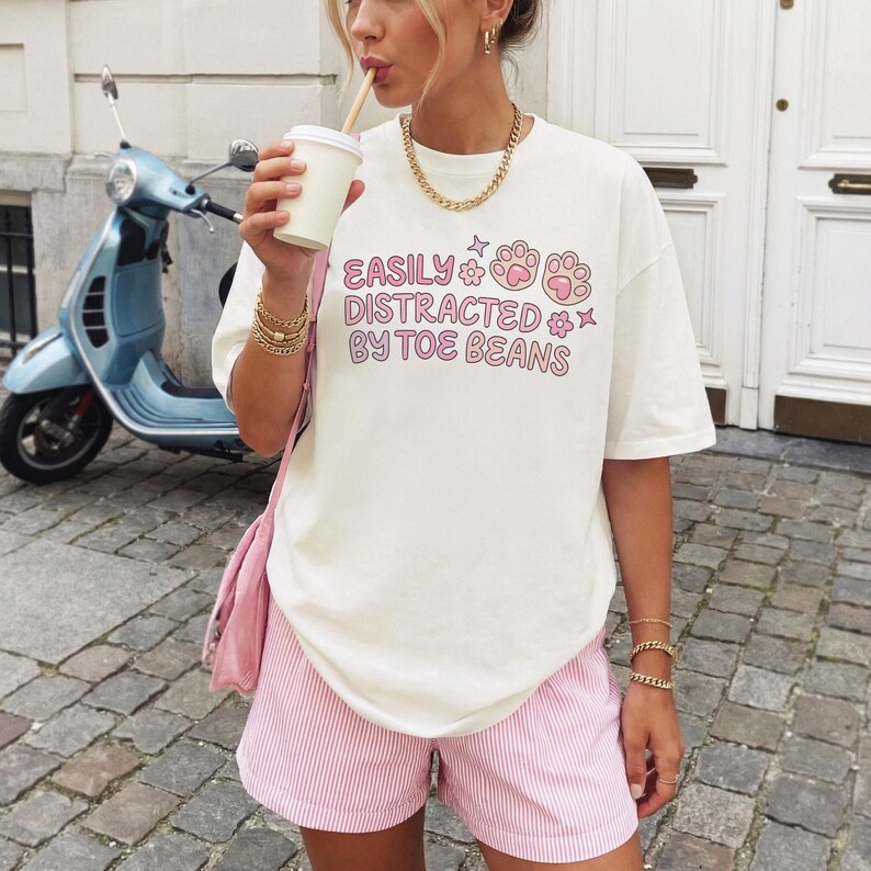 May include: An off-white short-sleeve t-shirt featuring the pink text "EASILY DISTRACTED BY TOE BEANS" alongside pink paw print and flower graphics. The shirt is paired with light pink and white pinstriped shorts and a pink shoulder bag.