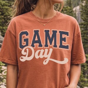 Op de afbeelding: Een roestkleurig T-shirt met korte mouwen met de tekst "GAME" in donkerblauwe blokletters met witte stippen, en het woord "Day" eronder in een groot, cr&egrave;mekleurig cursief lettertype. Het T-shirt heeft een ronde halslijn.