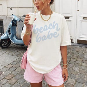 May include: Cream-colored t-shirt with the text "beach babe" in light blue, paired with pink and white striped shorts. A gold chain necklace and bracelets complete the look. A light blue scooter is in the background.