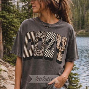 Op de afbeelding: Een antracietgrijs T-shirt met korte mouwen en het woord COZY in grote blokletters. Elke letter heeft een eigen patroon: verticale strepen, luipaardprint, een zwart-wit dambordpatroon en stippen met een slagschaduw.