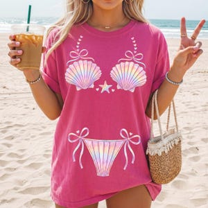 Può includere: Una t-shirt a maniche corte di colore rosa acceso con una stampa grafica di un bikini composto da due conchiglie iridescenti e uno slip abbinato. Il design include piccoli fiocchi, una stella marina centrale e dei cuoricini.