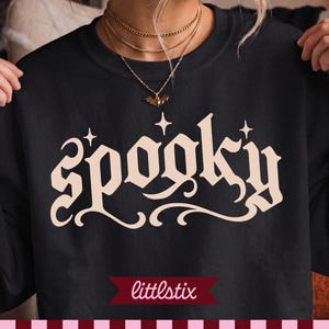 Puede incluir: Una sudadera negra de cuello redondo con la palabra SPOOKY impresa en una fuente gótica grande de color crema en el pecho. El diseño incluye pequeños detalles de estrellas sobre las letras y un colgante de murciélago en una cadena dorada.
