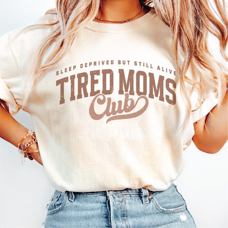 Puede incluir: Camiseta color crema con el texto "SLEEP DEPRIVED BUT STILL ALIVE TIRED MOMS Club" en letras marrones. La camiseta tiene un corte holgado y est&aacute; hecha de un material suave. El texto est&aacute; en negrita, estilo vintage.