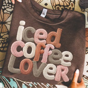 May include: A brown sweatshirt with the words "iced coffee lover" in a knitted, multi-colored font. The letters are in shades of white, pink, brown, and gray. The sweatshirt is held up by a person.