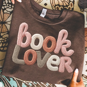 May include: A brown sweatshirt featuring the text "book lover" in a 3D knitted design. The letters are in shades of pink, white, brown, and grey. The sweatshirt is displayed on a patterned surface.