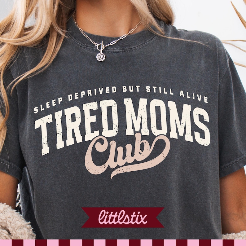 May include: A charcoal gray short-sleeve t-shirt featuring the white text "SLEEP DEPRIVED BUT STILL ALIVE" above the large white and light pink text "TIRED MOMS CLUB" in a distressed font style. The shirt has a crew neckline and a relaxed fit.