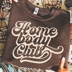 Peut inclure: Un sweat-shirt marron fonc&eacute; avec le texte "Home Body Club" en police cr&egrave;me de style r&eacute;tro. Le sweat-shirt est pli&eacute; et tenu par des mains, avec un fond &agrave; motifs visible.
