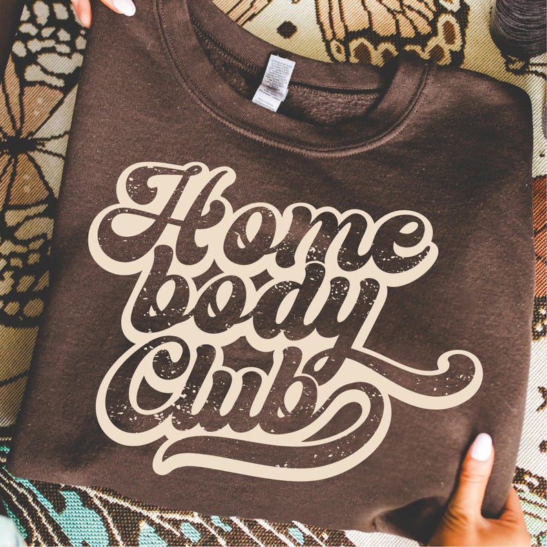 Peut inclure: Un sweat-shirt marron fonc&eacute; avec le texte "Home Body Club" en police cr&egrave;me de style r&eacute;tro. Le sweat-shirt est pli&eacute; et tenu par des mains, avec un fond &agrave; motifs visible.