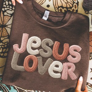 May include: A dark brown crewneck sweatshirt featuring the text "JESUS LOVER" in a three-dimensional, knit-textured font. The letters are rendered in various muted earth tones, including shades of cream, beige, tan, and dusty rose, creating a tactile appearance.
