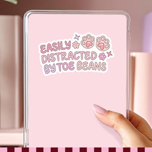 May include: A rectangular tablet case with a light pink back featuring a white sticker. The sticker displays the text EASILY DISTRACTED BY TOE BEANS in pink letters, accompanied by two pink cat paw prints, two small flowers, and two four-pointed stars.