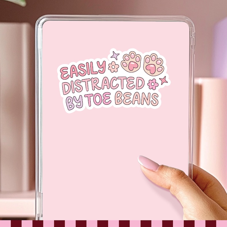 May include: A rectangular tablet case with a light pink back featuring a white sticker. The sticker displays the text EASILY DISTRACTED BY TOE BEANS in pink letters, accompanied by two pink cat paw prints, two small flowers, and two four-pointed stars.