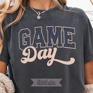 Op de afbeelding: Een antracietgrijs T-shirt met korte mouwen met het woord GAME in donkerblauwe blokletters met witte stippen, en het woord Day in lichtbeige cursief schrift. Een kleine grijze banner met de tekst littlestix staat onderaan.