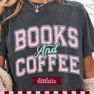 May include: A charcoal gray short-sleeve t-shirt featuring the text BOOKS And COFFEE in a distressed pink and light blue varsity-style font. The word BOOKS is on the top line, And is in script in the middle, and COFFEE is on the bottom line.
