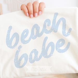May include: Cream-colored tote bag with the words "beach babe" in light blue, distressed lettering. The bag is held up by a hand with pink nail polish. The brand name "littlestix" is at the bottom.