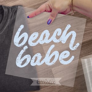 May include: A gray t-shirt with a transparent transfer sheet featuring the words "beach babe" in light blue, distressed font. The transfer sheet is being held over the shirt, with the brand name "littlestix" at the bottom.