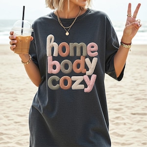 May include: Dark gray t-shirt with the words "home body cozy" in a textured, multi-colored design. The shirt is oversized and appears to be made of a soft material. The image is taken on a beach.