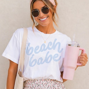 May include: White t-shirt with the text "beach babe" in light blue script. A pink insulated tumbler with a handle and straw is held in the hand. The person is wearing sunglasses and a pink headband.