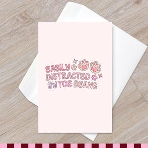 May include: A light pink greeting card featuring the text EASILY DISTRACTED BY TOE BEANS in a bubbly font, accompanied by two illustrated cat paw pads, small flowers, and star shapes, resting on top of a plain white envelope.