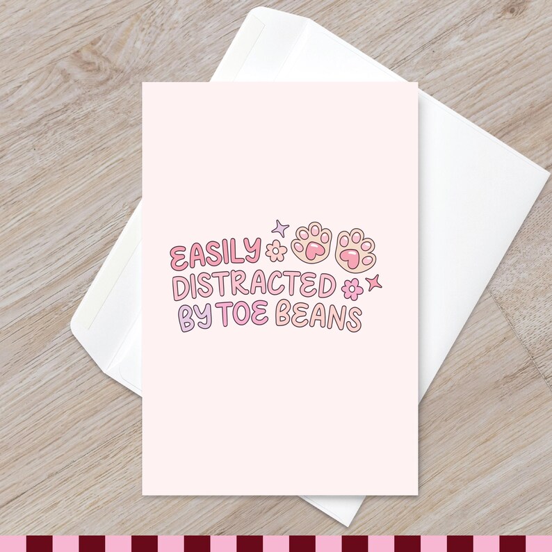 May include: A light pink greeting card featuring the text EASILY DISTRACTED BY TOE BEANS in a bubbly font, accompanied by two illustrated cat paw pads, small flowers, and star shapes, resting on top of a plain white envelope.