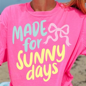 May include: A vibrant pink t-shirt featuring the text "MADE for SUNNY days" in light blue and yellow, with a light pink bow illustration. The casual top is ideal for sunny days and warm weather.