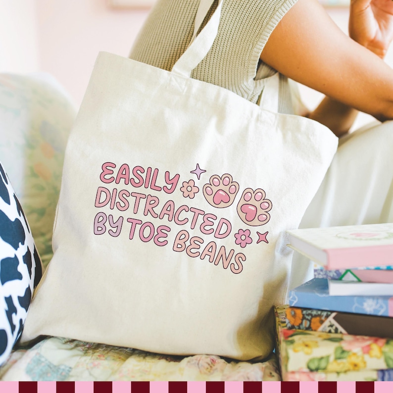 May include: A cream-colored canvas tote bag featuring the text "EASILY DISTRACTED BY TOE BEANS" in a pink, rounded font. The design includes two pink cat paw prints, small flower icons, and star shapes printed on the front of the fabric bag.
