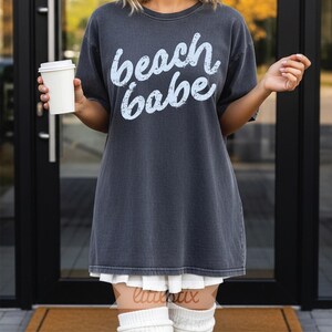 May include: Dark gray oversized t-shirt with the words "beach babe" in light blue script. The shirt is worn with a white pleated skirt and white knee-high socks. A person is holding a white paper coffee cup.