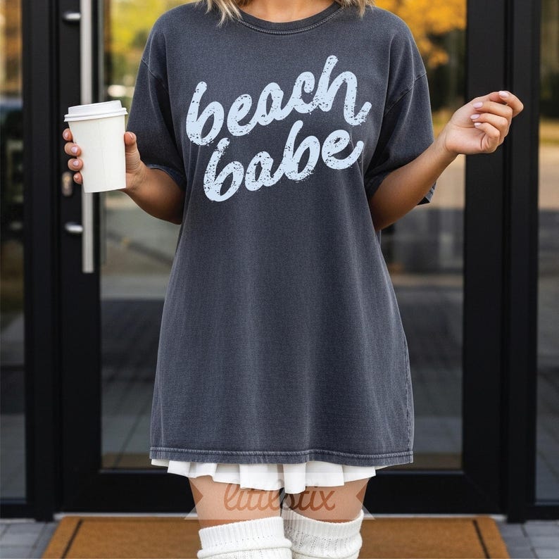 May include: Dark gray oversized t-shirt with the words "beach babe" in light blue script. The shirt is worn with a white pleated skirt and white knee-high socks. A person is holding a white paper coffee cup.