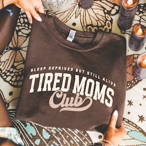 Puede incluir: Una sudadera marr&oacute;n con el texto "SLEEP DEPRIVED BUT STILL ALIVE TIRED MOMS Club" en letras blancas y beige. La sudadera es sostenida por manos, con velas y una tela estampada en el fondo.