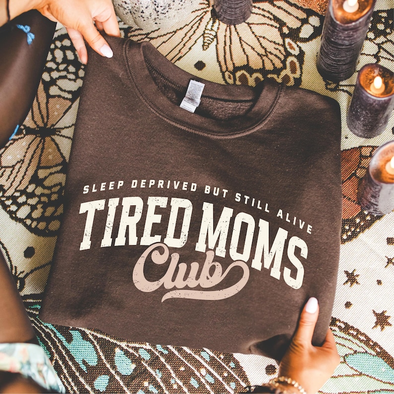 Puede incluir: Una sudadera marr&oacute;n con el texto "SLEEP DEPRIVED BUT STILL ALIVE TIRED MOMS Club" en letras blancas y beige. La sudadera es sostenida por manos, con velas y una tela estampada en el fondo.
