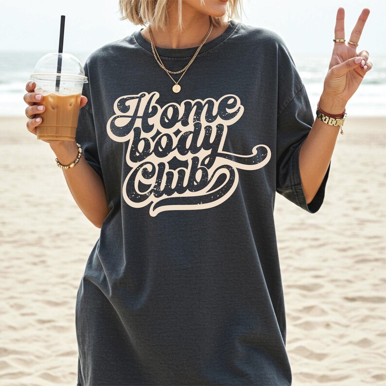 Peut inclure: T-shirt gris fonc&eacute; avec l'inscription "Home Body Club" dans une police r&eacute;tro. La personne porte le t-shirt et tient un caf&eacute; glac&eacute; et fait un signe de paix. L'arri&egrave;re-plan est une plage de sable avec l'oc&eacute;an au loin.