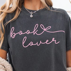 May include: Dark gray t-shirt with the pink text "book lover" in a cursive font. The shirt is a comfortable, casual style. A silver necklace with a pendant is visible at the neckline.
