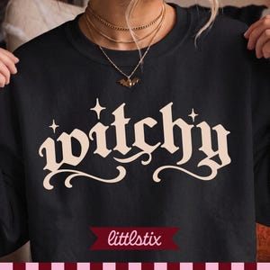 Puede incluir: Una sudadera negra de cuello redondo con la palabra "witchy" impresa en una fuente gótica grande de color crema en el pecho. El texto está acentuado con pequeños iconos de estrellas de cuatro puntas sobre las letras. Tela negra sólida.