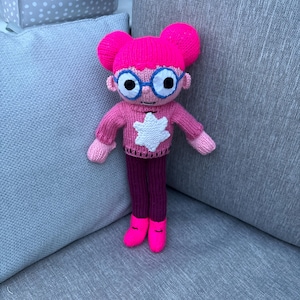 May include: A knitted doll with pink hair buns, blue-rimmed glasses, a pink sweater with a white star, purple trousers, and pink shoes. The doll has its arms outstretched.
