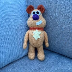 May include: A handmade, knitted plush toy in the shape of a cartoon character. The toy is primarily peach-colored with brown ears, a purple nose, and a white patch. It has an orange scarf and a white star on its chest.