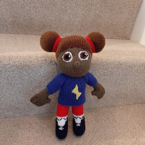 May include: A knitted doll with brown skin, brown hair in two pigtails, a blue sweater with a yellow lightning bolt, red trousers, and black shoes.
