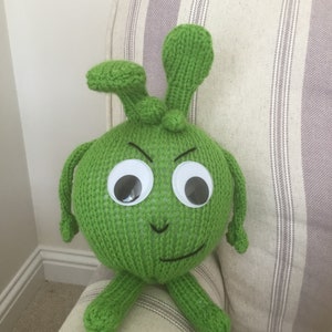 May include: Green knitted plush toy with large white eyes and a frowning expression. The toy has two long arms and two short legs.