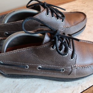 May include: A pair of brown leather boat shoes with black laces. The shoes have a rubber sole and are laced up the front.