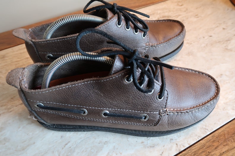 May include: A pair of brown leather boat shoes with black laces. The shoes have a rubber sole and are laced up the front.