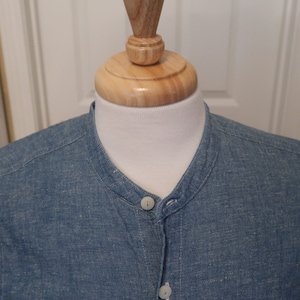 May include: A blue denim shirt with a mandarin collar and a single button closure. The shirt has a textured, slightly rough surface.