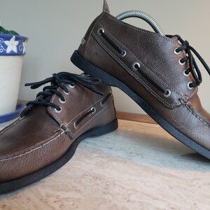 May include: A pair of brown leather boat shoes with black laces. The shoes have a moc toe design and a rubber sole.