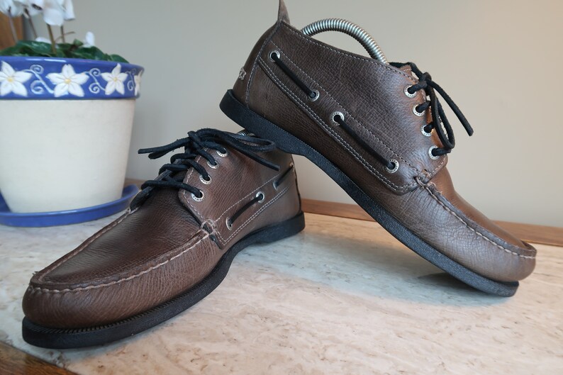 May include: A pair of brown leather boat shoes with black laces. The shoes have a moc toe design and a rubber sole.