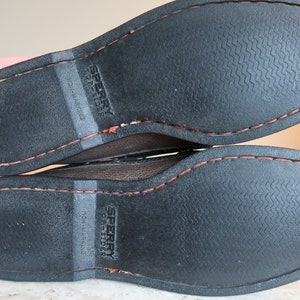 May include: Close-up of the bottom of a pair of brown leather Sperry Top-Sider boat shoes. The shoes have a black rubber sole with a textured pattern and a red stitching detail around the edge.