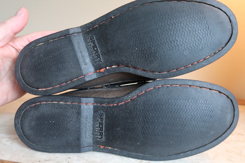 May include: Close-up of the bottom of a pair of brown leather Sperry Top-Sider boat shoes. The shoes have a black rubber sole with a textured pattern and a red stitching detail around the edge.